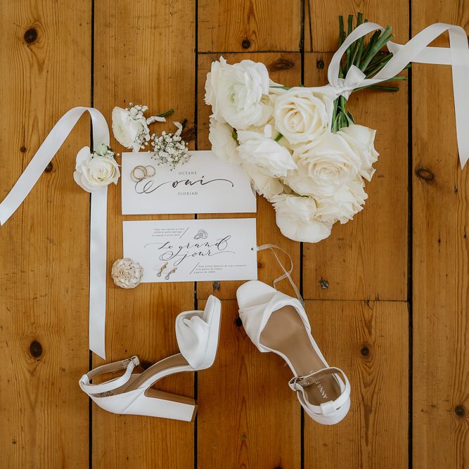 Les Craneuses, agence de wedding planner dans les Hauts-De-France Chaussures blanches, fleurs, enveloppes et rubans sur un sol en bois.
